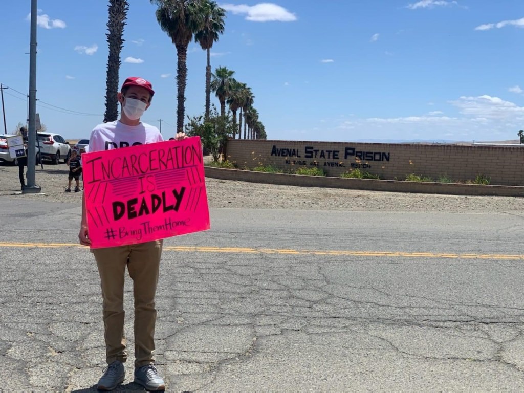 man holds bright pink sign protesting covid cases in prison. sign reads "incarceration is deadly"