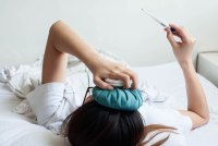 A young woman with ice pack on her head is checking on her temperature using a digital thermometer while lying on her back