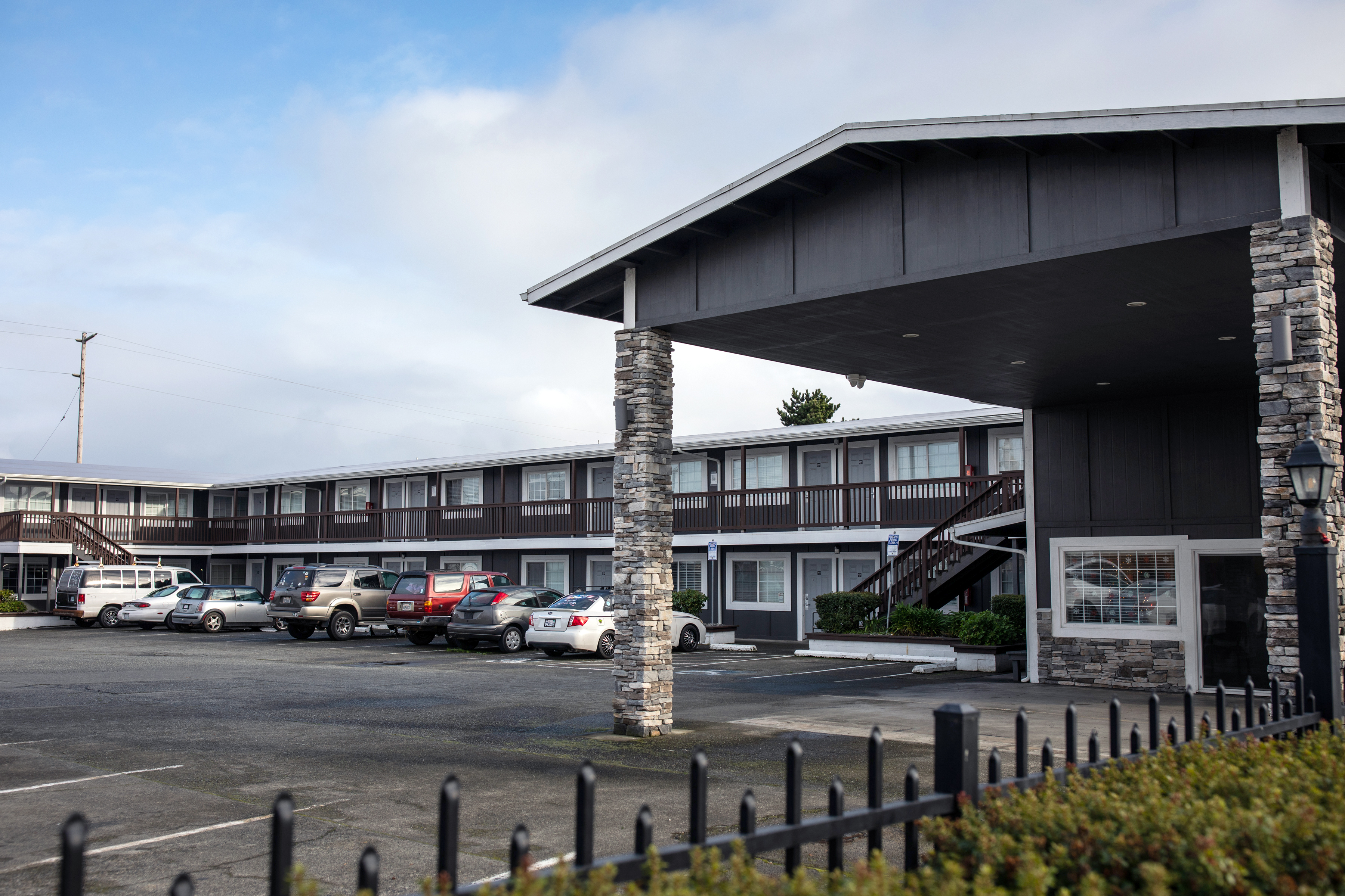 A variety of cars are parked in the parking lot of The Legacy, a housing center for those who are homeless in Del Norte County converted from an old motel. The renovated building is painted a dark gray.