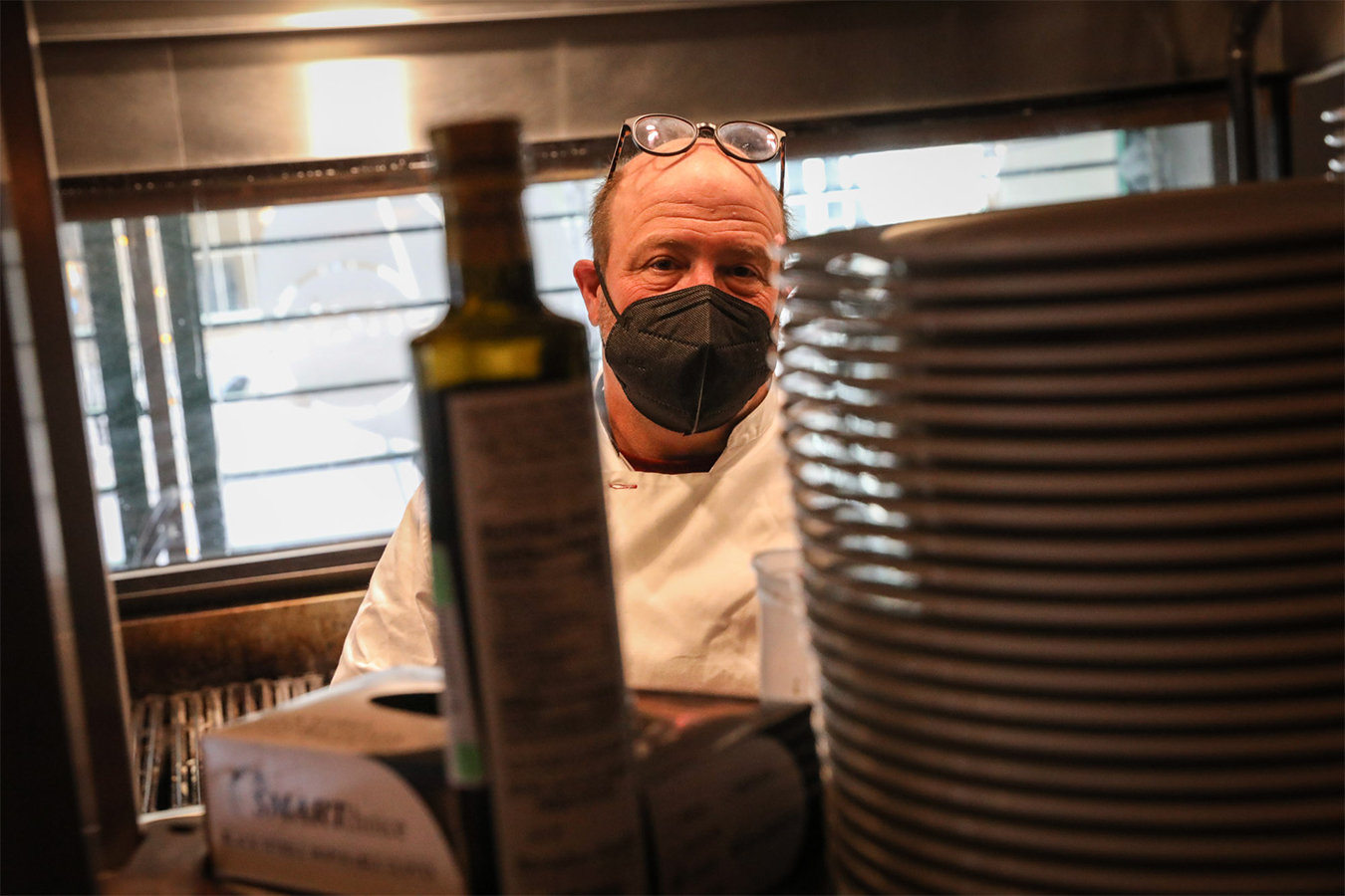 Frank Bonanno is seen from behind a stack of plates and a bottle of olive oil.  He is in the kitchen of his restaurant, French 75.
