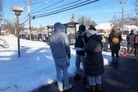 A long line of people waiting for a covid test stretches along the sidewalk of a community. A few inches of snow has been shoveled away to clear a path, and everyone is wearing thick winter coats.