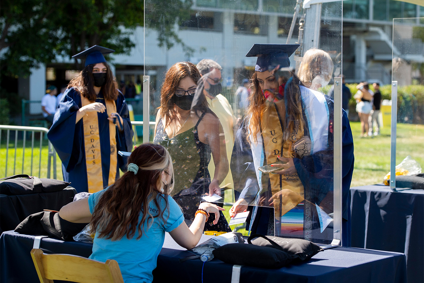 Two women hold out their vaccine cards to a person at an outdoor booth behind a plastic face shield. The woman on the right is wearing a blue graduation cap and gown, another woman stands to her left, in normal clothes.