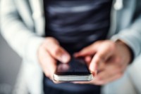 A closeup photo shows a man holding a smartphone in his hands. The shot is tightly focused on his phone, the rest of the photo is blurred.