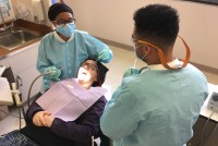 Mary Ashlee Tosh lies in a dental chair while Dr. Ratrice Jackson sits to her side, holding dental tools in both of hands. A man is seen in the foreground in the left of the frame.