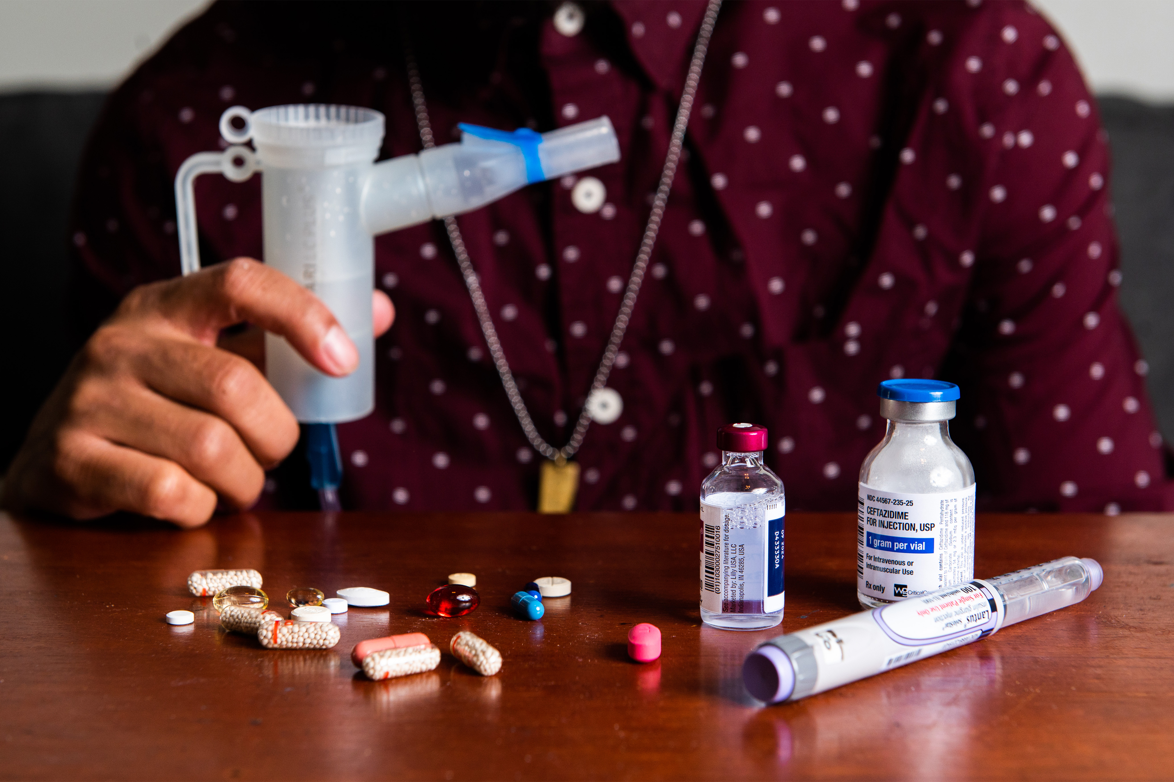 یک نمای نزدیک، دست راست نیکلاس کلی را نشان می‌دهد که یک دستگاه اتومایزر با مجموعه‌ای از قرص‌ها و ویال‌های دارویی را روی میزی در مقابل او نگه داشته است.
