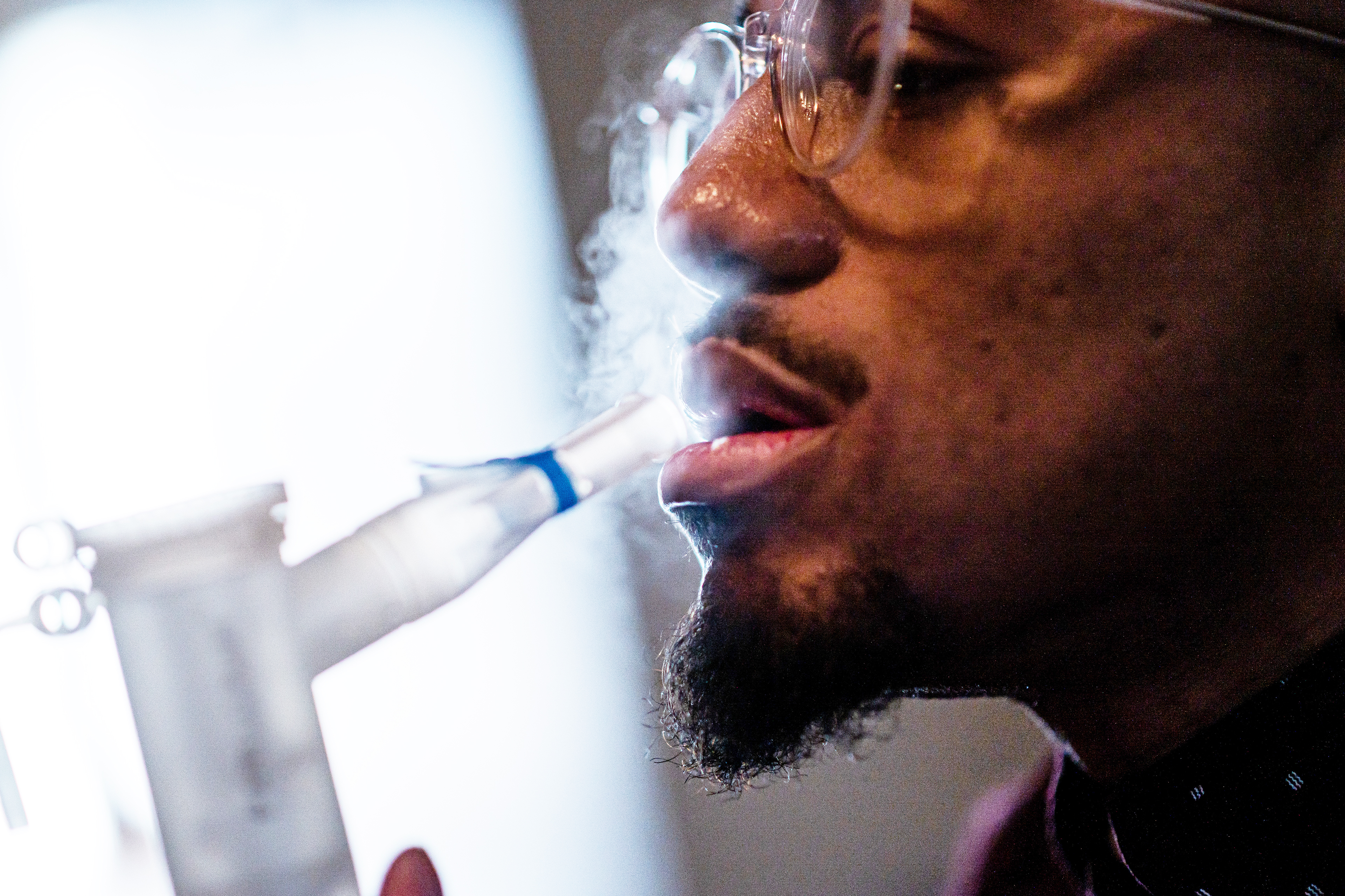 نبولایزر دارویی را اسپری می کند که نیکلاس کلی، که در تصویر نمایه سمت راست نشان داده شده است، از طریق یک قطعه دهانی تنفس می کند.  پنجره ای در پس زمینه ظاهر می شود که سایه ای دراماتیک ایجاد می کند.