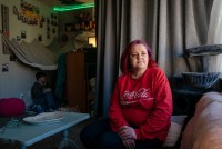 Stacy Whitford sits in her house on the right, facing a window. Her son sits on the floor in the corner on the left.