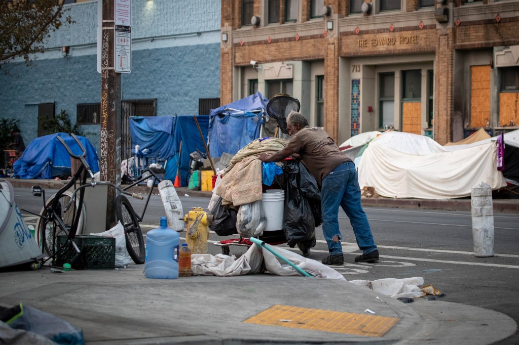 A man is seen pushing a full cart of belongings through the street. Tents and makeshift shelters are seen in the background behind him.