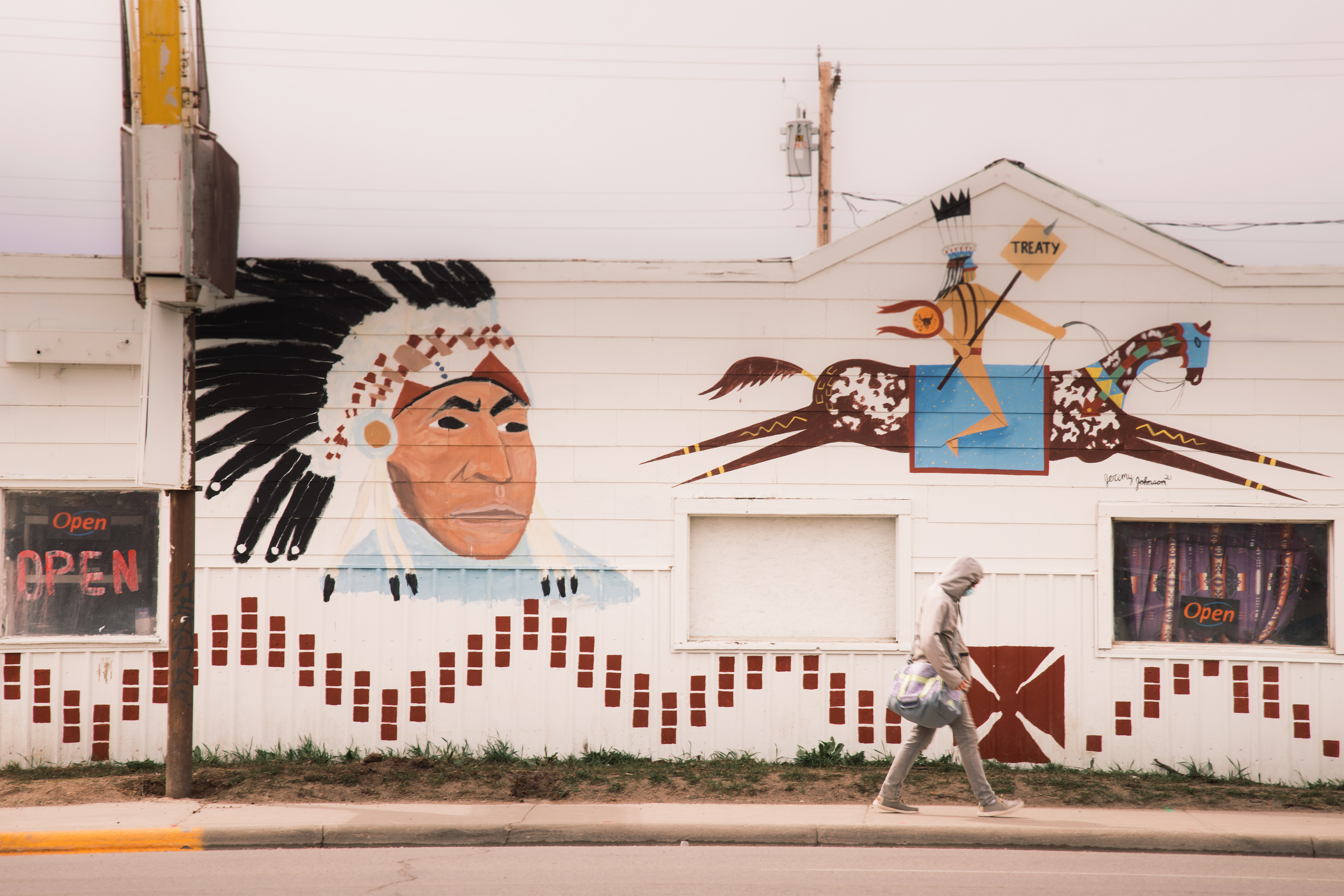 A person is seen walking on the sidewalk by a building in Browning, Montana. The side of the building is painted with an image of a Native American man wearing a war bonnet. Another painting shows a Native American on horseback holding a spear piercing through a sign that reads, "TREATY."