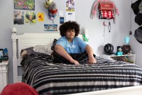 A wide shot photo shows Cameron Wright sitting in his bed at home in Denton, Texas. Posters and trinkets hang on the wall behind him.
