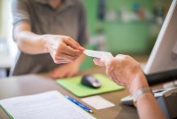 A closeup photo shows a woman handing over an insurance card to someone behind a counter.