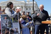 Senator Chuck Schumer is seen standing at a podium with a microphone, talking to a crowd at a press conference. People hold signs and are seen wearing veteran's hats around him.