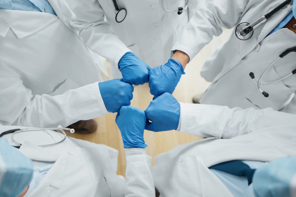 Five doctors in doctors in lab coats and blue gloves fist-bump.