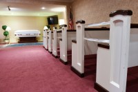 A wide shot shows rows of pews in a funeral home leading up to casket. Some of the pews are cordoned off.