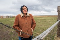 Marla Ollinger is seen standing behind a fence on her ranch in Browning, Montana. She is wearing a jacket with her hands in her pockets.