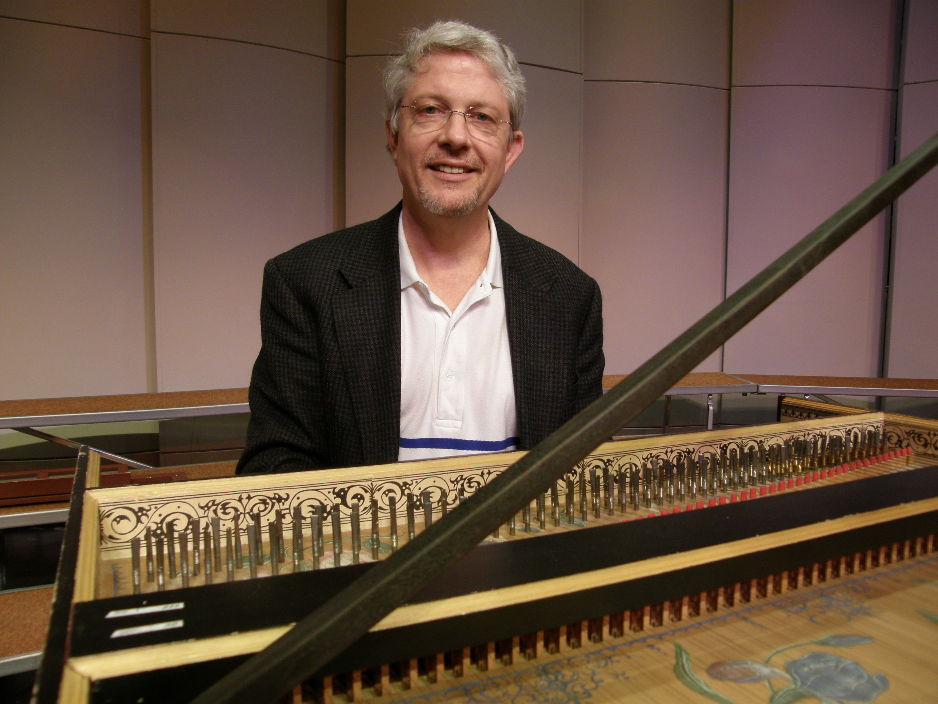 Richard Gard is seen smiling for a photo, sitting in front of a harpsichord. 
