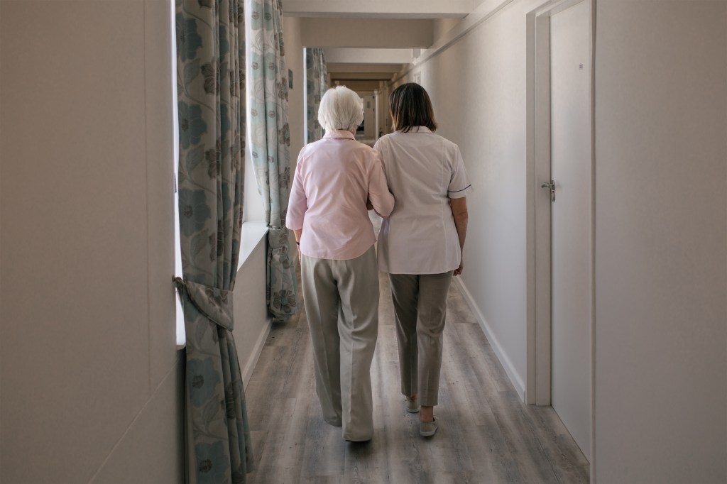 A photo shows a nurse walking an elderly woman down a hallway in a nursing home. The two are seen from behind.