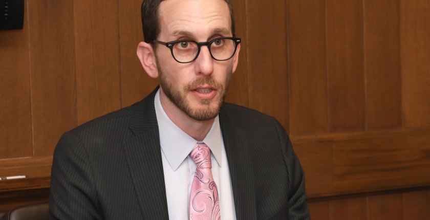 A man in a dark suit and light pink tie wearing glasses speaks.