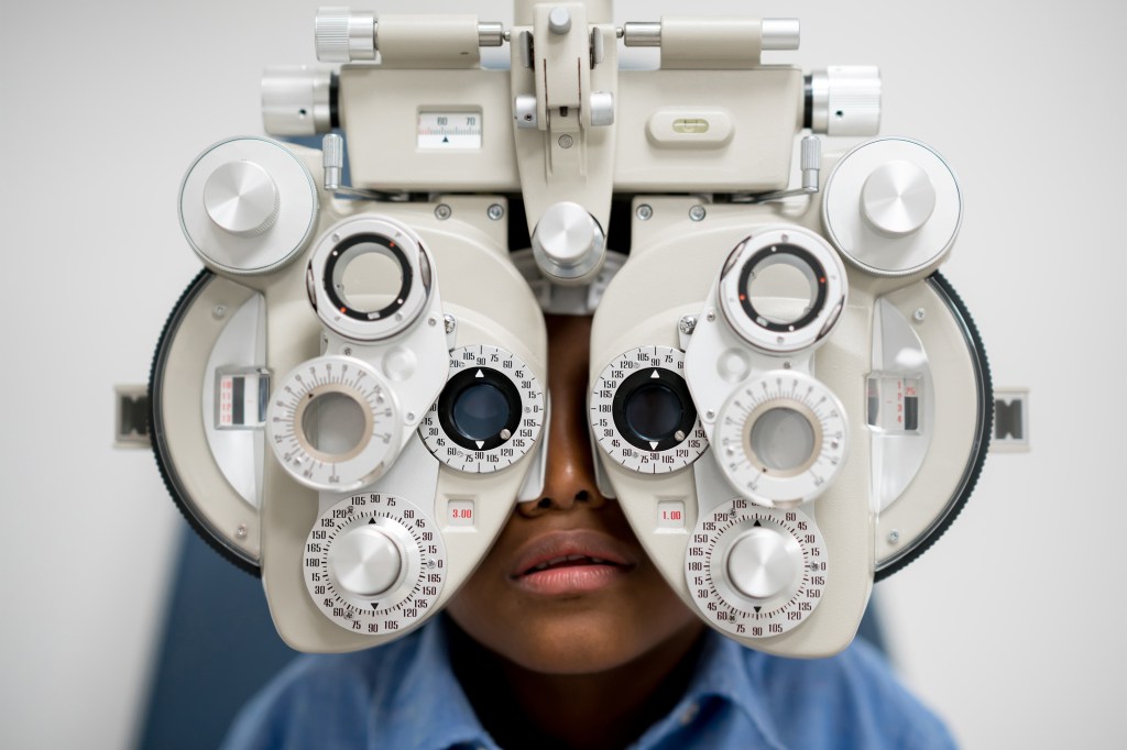 A closeup photo shows a child viewed from the front getting an eye exam with a phoropter. The large medical instrument obscures most of the child's face from view.