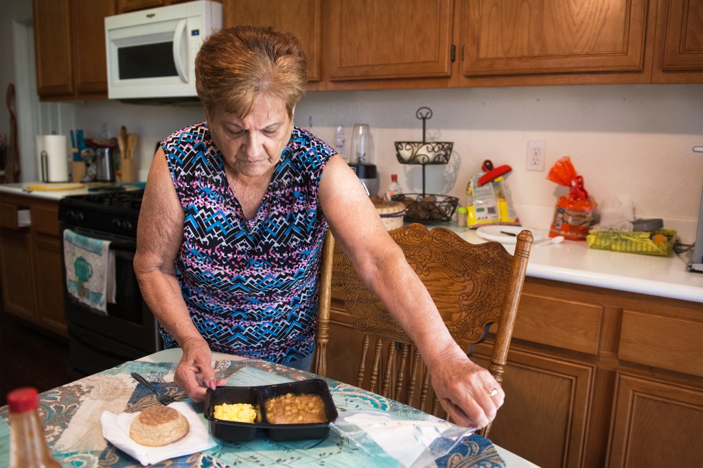 A photo shows Frances De Los Santos in her kitchen peeling off the wrapper of a prepared meal in a frozen food tray. The meal was delivered to her home thanks to a new Medi-Cal program.