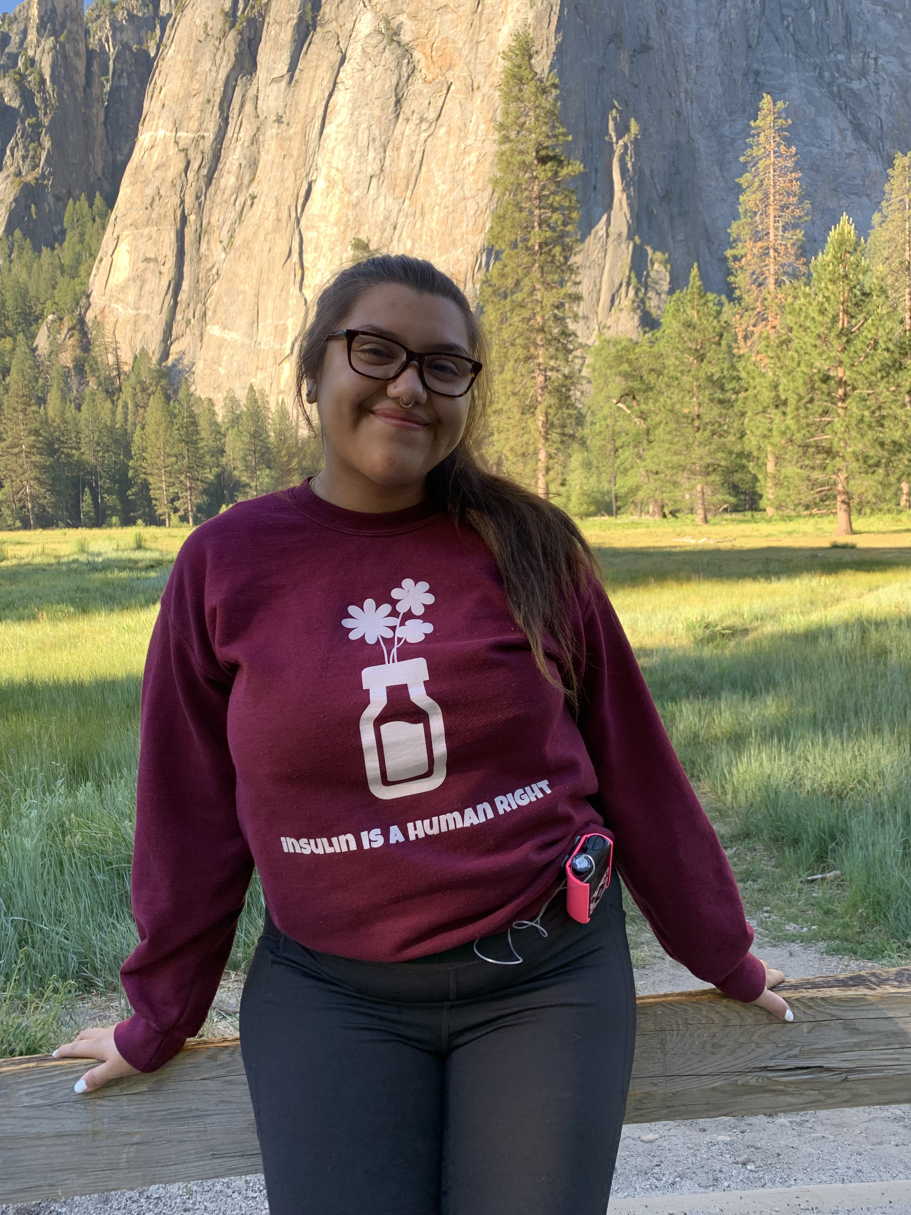 Sabrina Caudillo is standing outdoors, smiling at the camera. She wears a maroon sweatshirt that reads, "INSULIN IS A HUMAN RIGHT" in white letters. Above the text is a design of flowers coming out of an insulin vial, also in white..