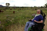 A photograph of Bruce Goguen sitting in his wheelchair outside, smiling at the camera. Beyond him is a sprawling pasture, where cows can be seen grazing.