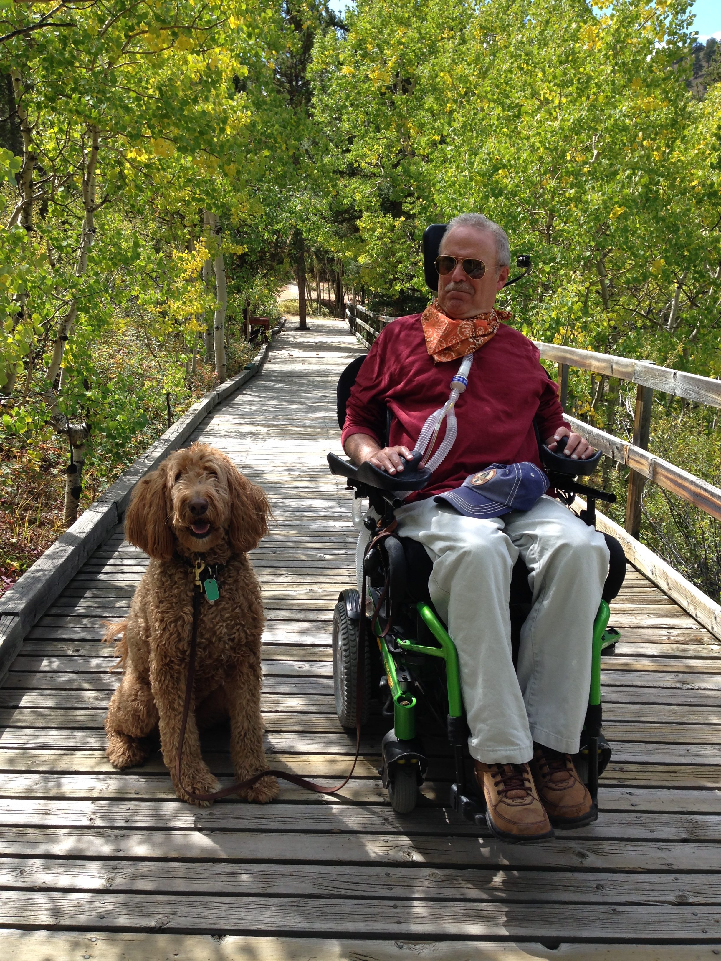Bruce Goguen is photographed in his wheelchair beside his dog. They are sitting outdoors, surrounded by nature.