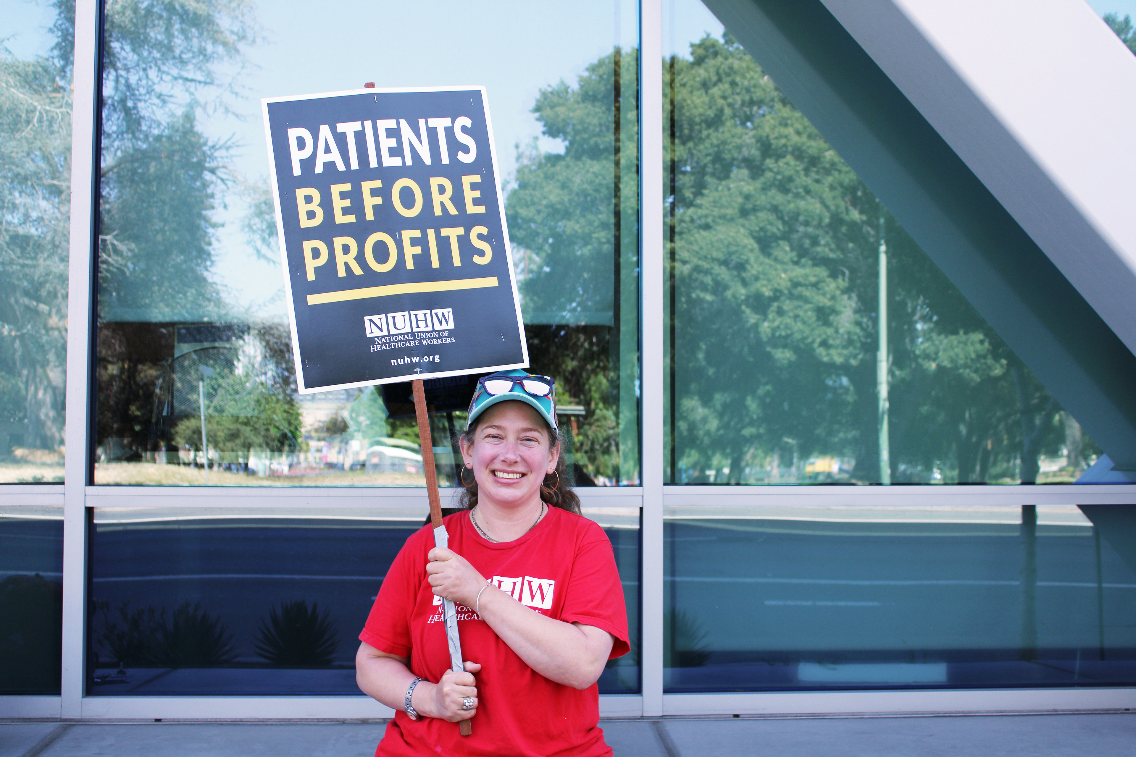A photo shows Alicia Moore standing outside Kaiser Permanente Hospital in Oakland, California.  She holds a sign that reads, "Patients before profits."