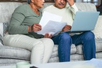 A photo shows an elderly couple sitting on a couch and looking over paperwork and a laptop together.
