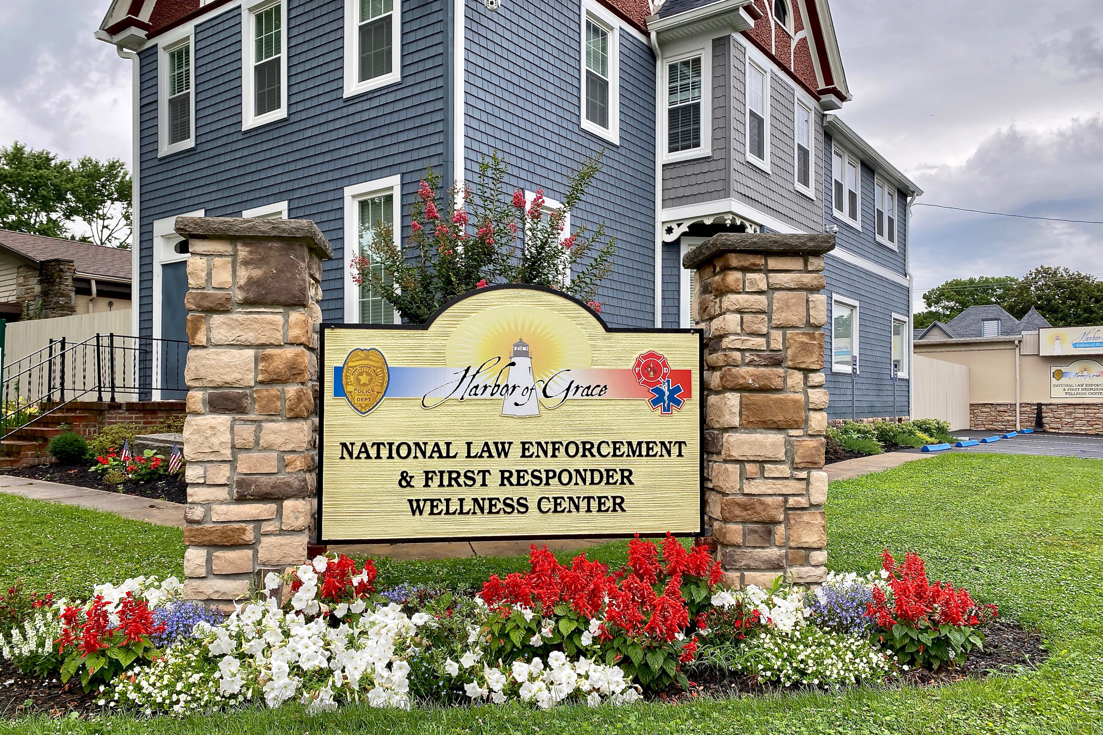 A photo shows a sign outside the Harbor of Grace Enhanced Recovery Center. The sign bears the name of the center and the words, "National Law Enforcement and First Responder Wellness Center," and is surrounded by flowers.