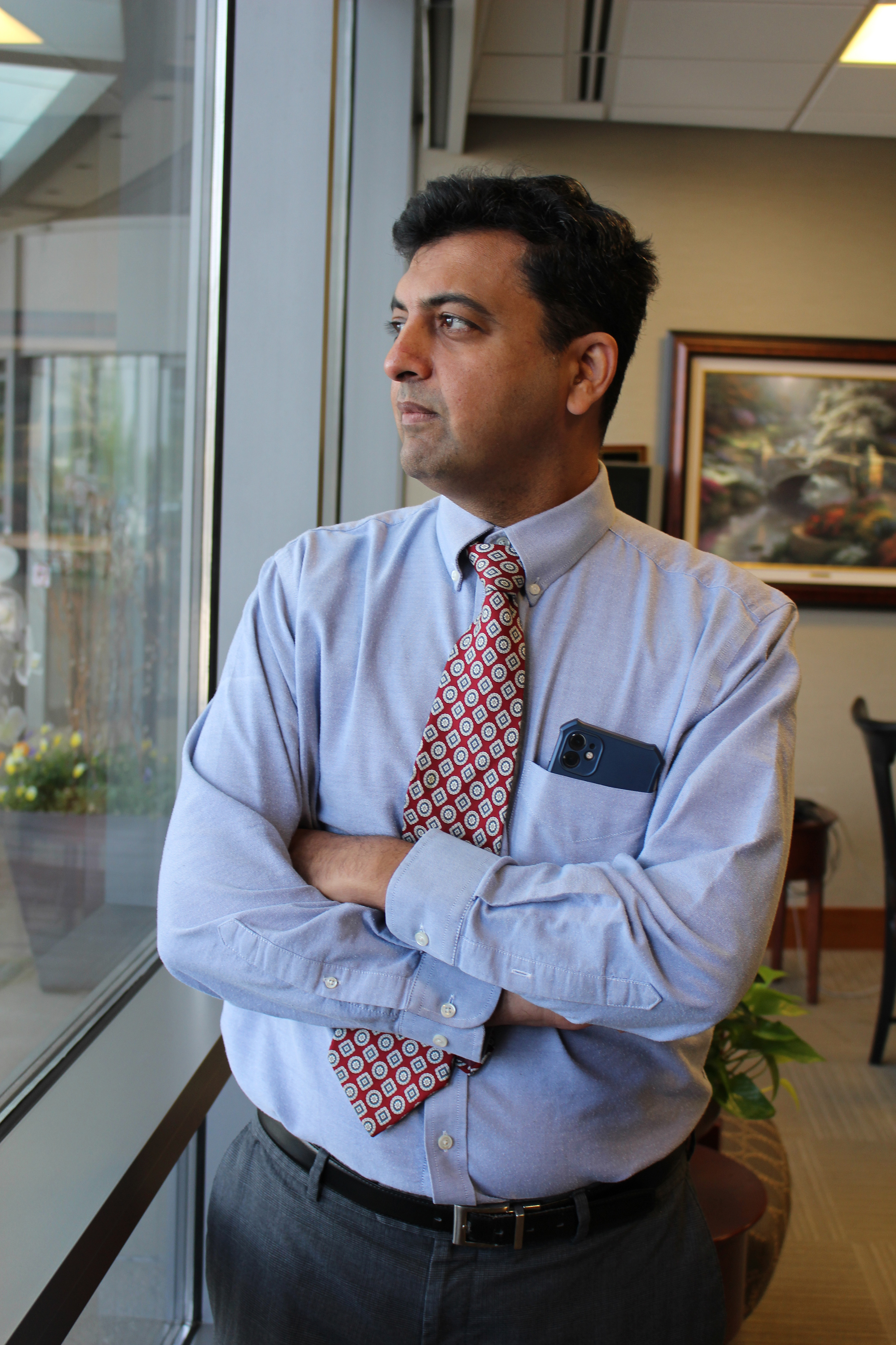 Dr. Bhavin Shah stands beside a window and is looking out with his arms crossed in front of his chest. He appears contemplative and wears a dress shirt and tie.