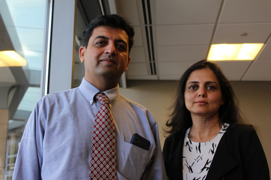 Dr. Bhavin Shah, who wears a dress shirt and tie, stands beside his wife, Sunita Kalsariya, who wears a black cardigan over a white patterned shirt. They are in an office room next to a window, and look towards the camera.