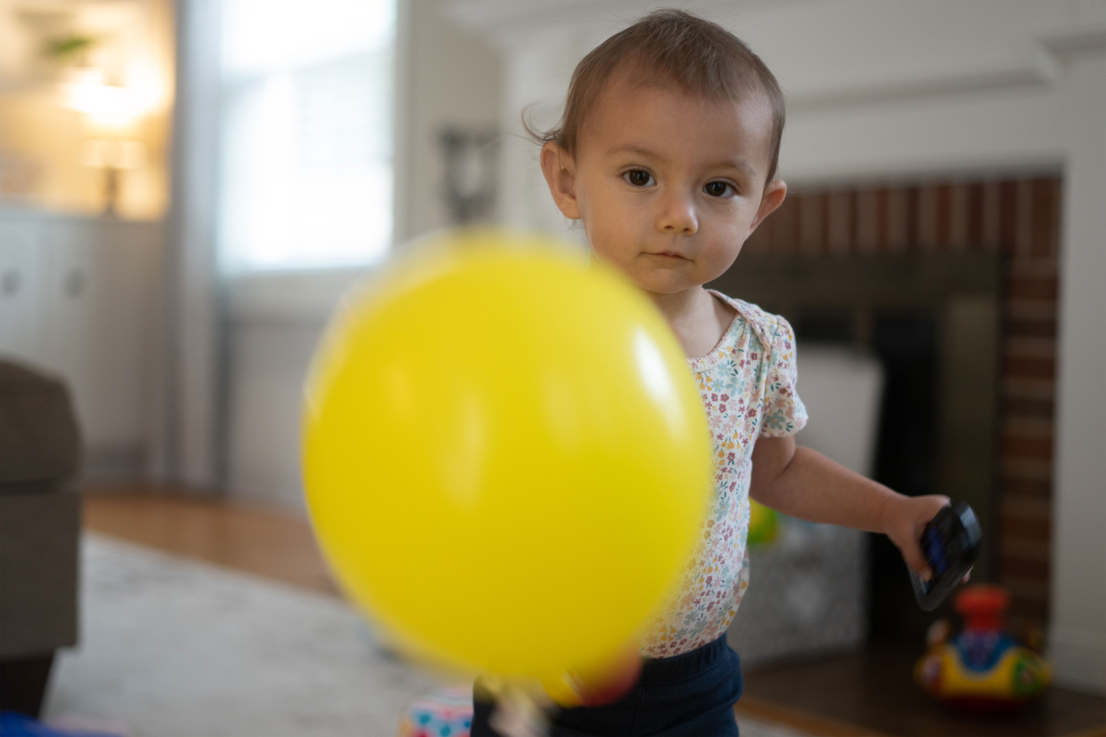 A girl about one year old looks at the camera and shows off a yellow balloon.