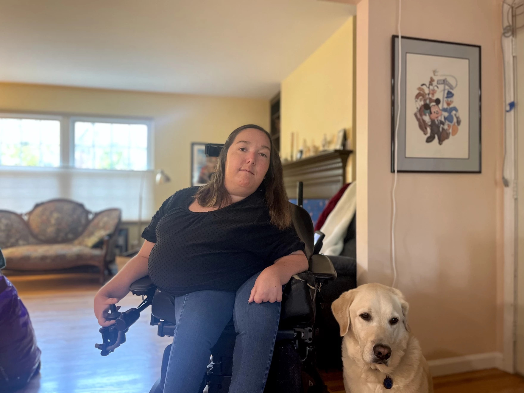 A photo shows Emily Munson in a wheelchair at home. A dog is sitting beside her.