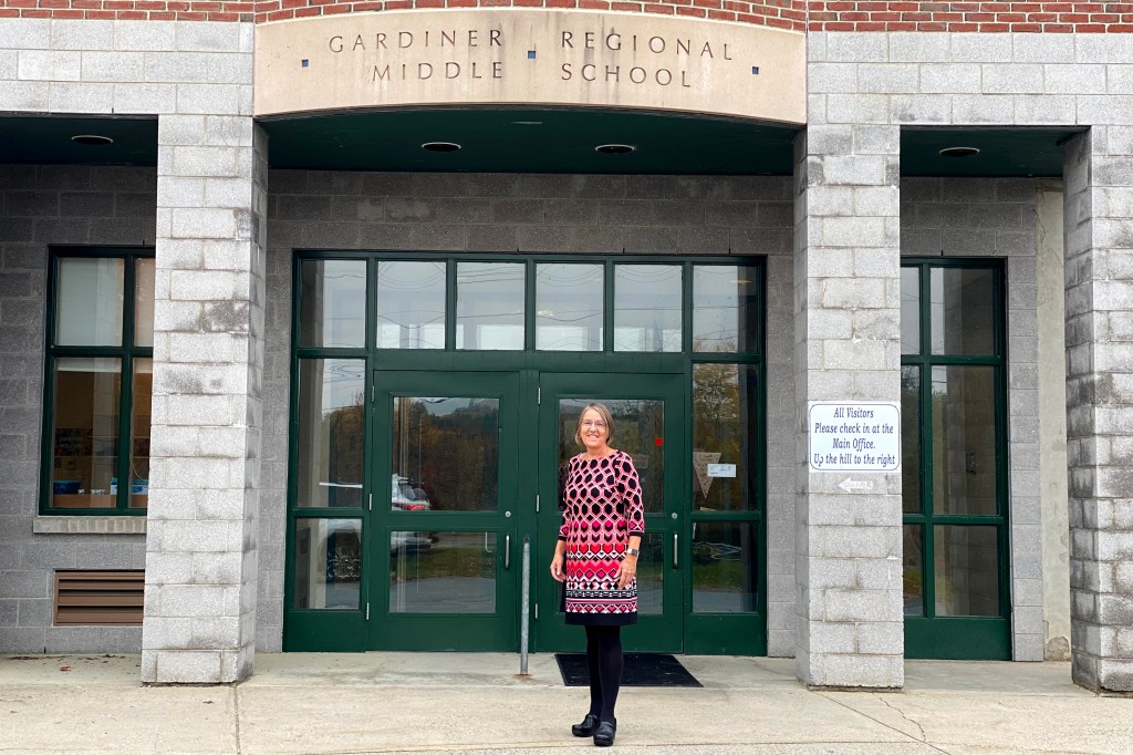 Patricia Hopkins, wearing a pink blouse, stands in front of the school building where she works. There are large green and glass doors behind her, and brick pillars to both sides.