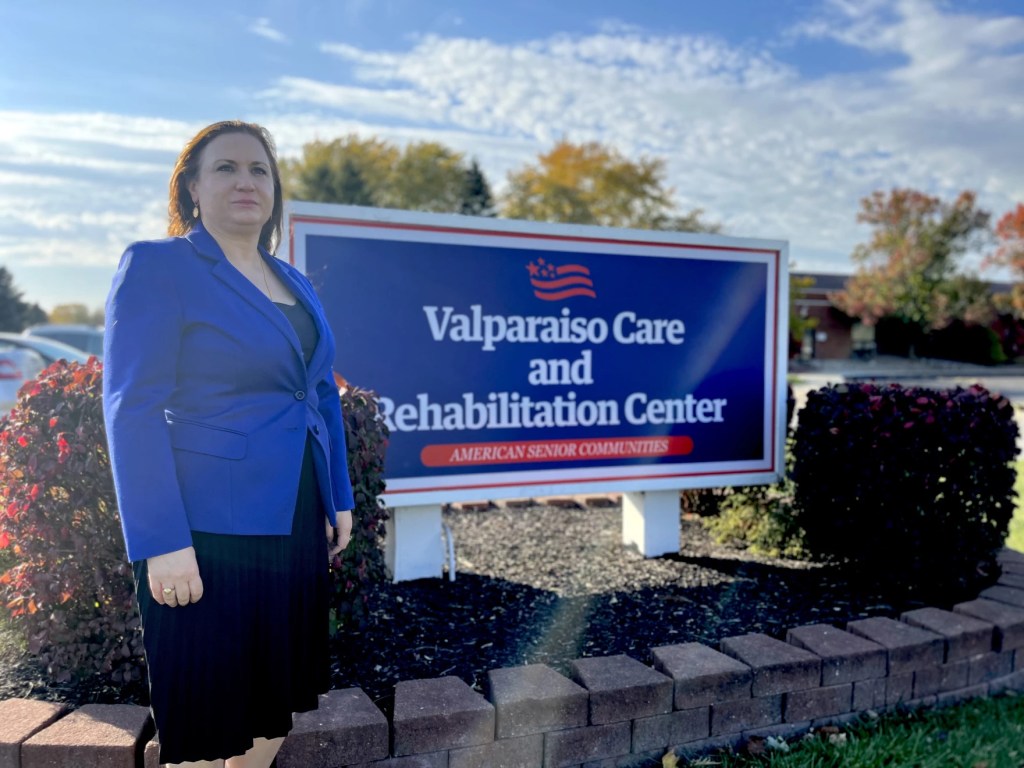 A photo shows Susie Talevski standing next to the sign outside the Valparaiso Care and Rehabilitation Center, the nursing home where her father resided.