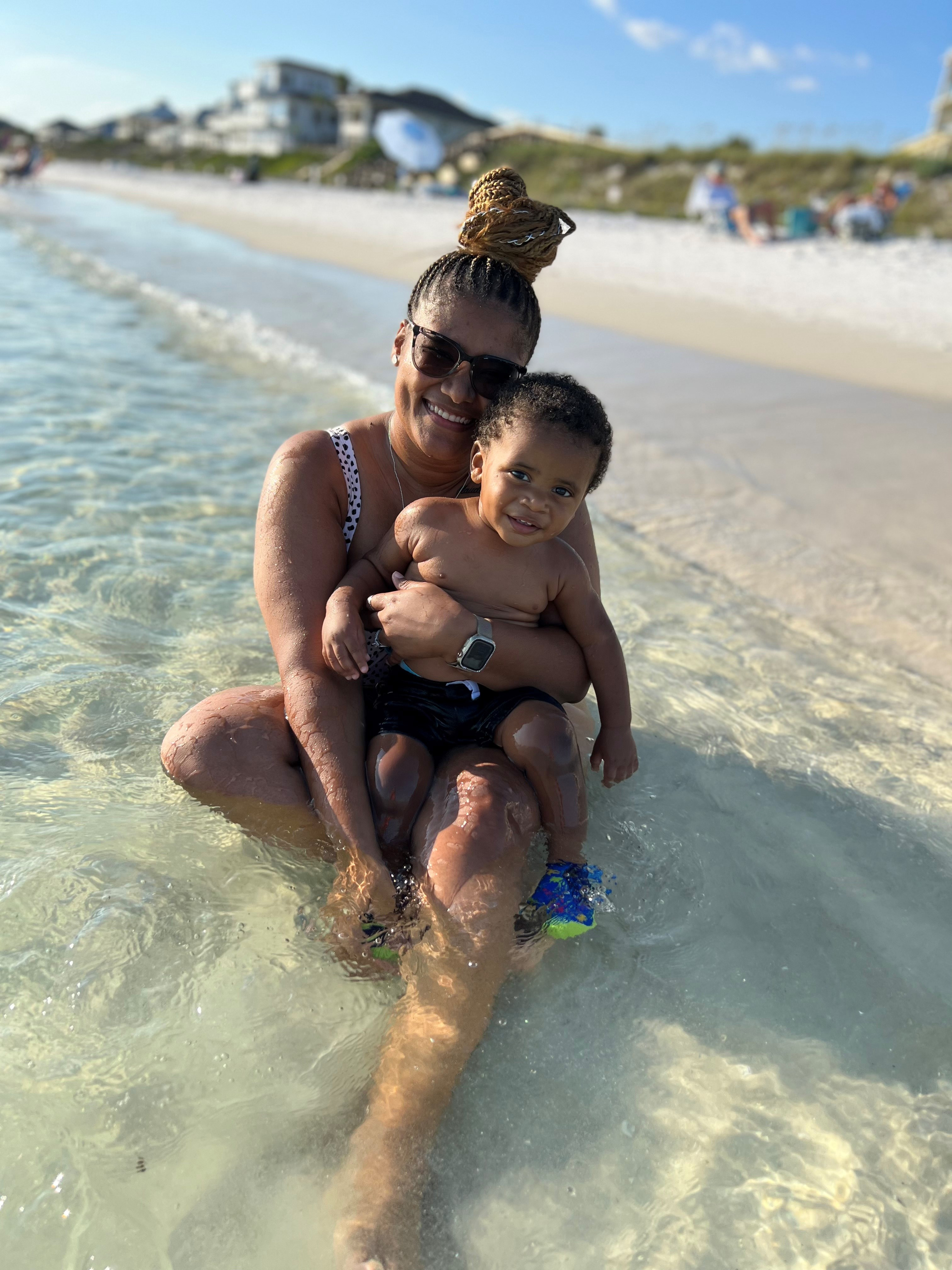 Mineka Furtch is sitting at the beach, in clear shallow water, with her 1-year old son in her lap.