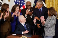 President Joe Biden, who is sitting at a desk, gives a pen to former President Barack Obama after signing an executive order aimed at strengthening the Affordable Care Act on April 5. Others stand around them, clapping.