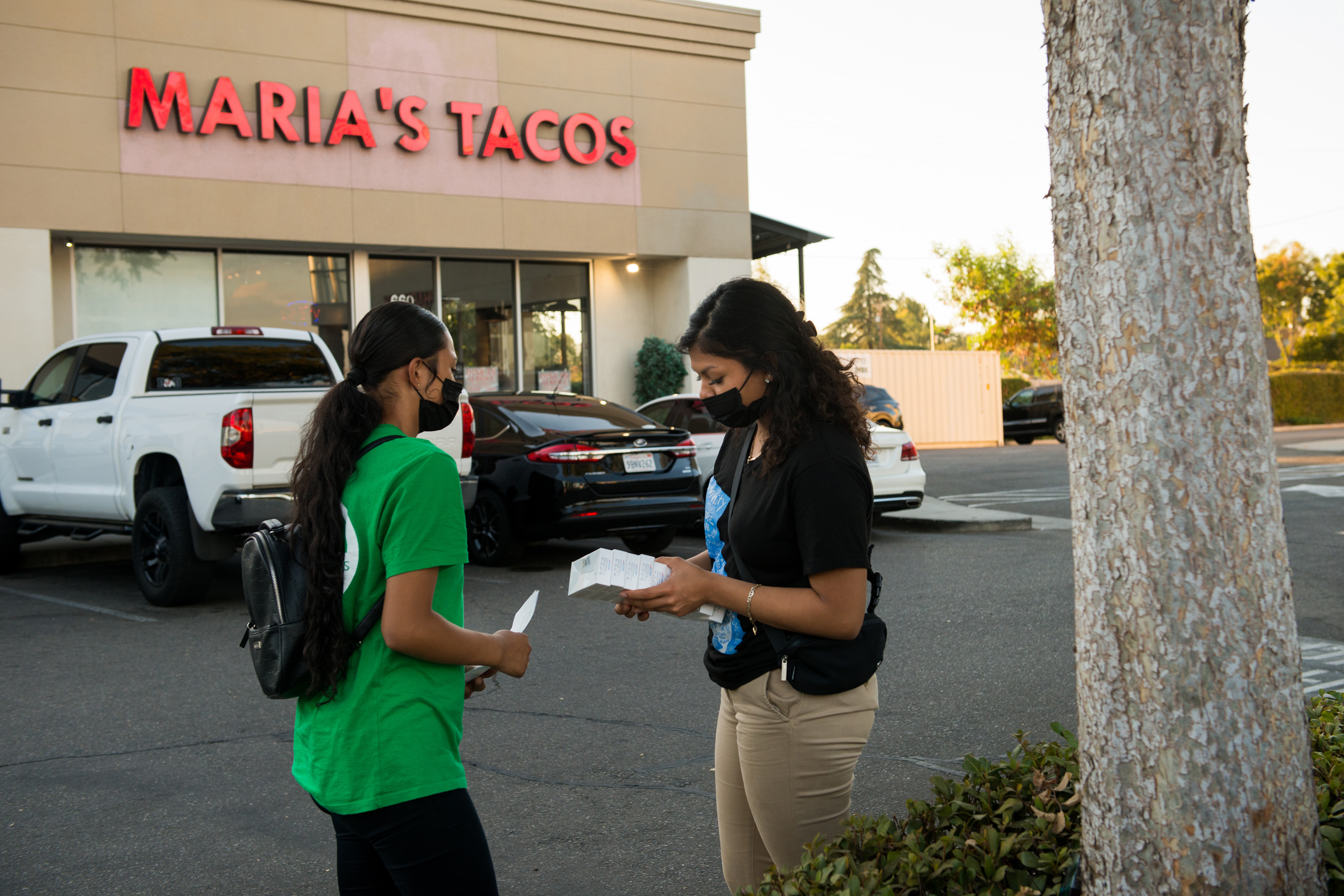 Melissa Lopez (left) and Alma Gallegos (right) get ready to distribute covid tests. They are standing in the parking lot in front of a building that says "Maria's Tacos."