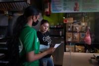 Melissa Lopez explains the covid testing process to a taco shop employee. They are in the back of the restaurant, and storage items are visible on the shelves behind them.