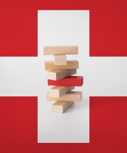 A stack of wooden blocks is seen surrounded by a medical cross. One of the wooden blocks in the center of the stack is painted red, indicating the tower may fall.