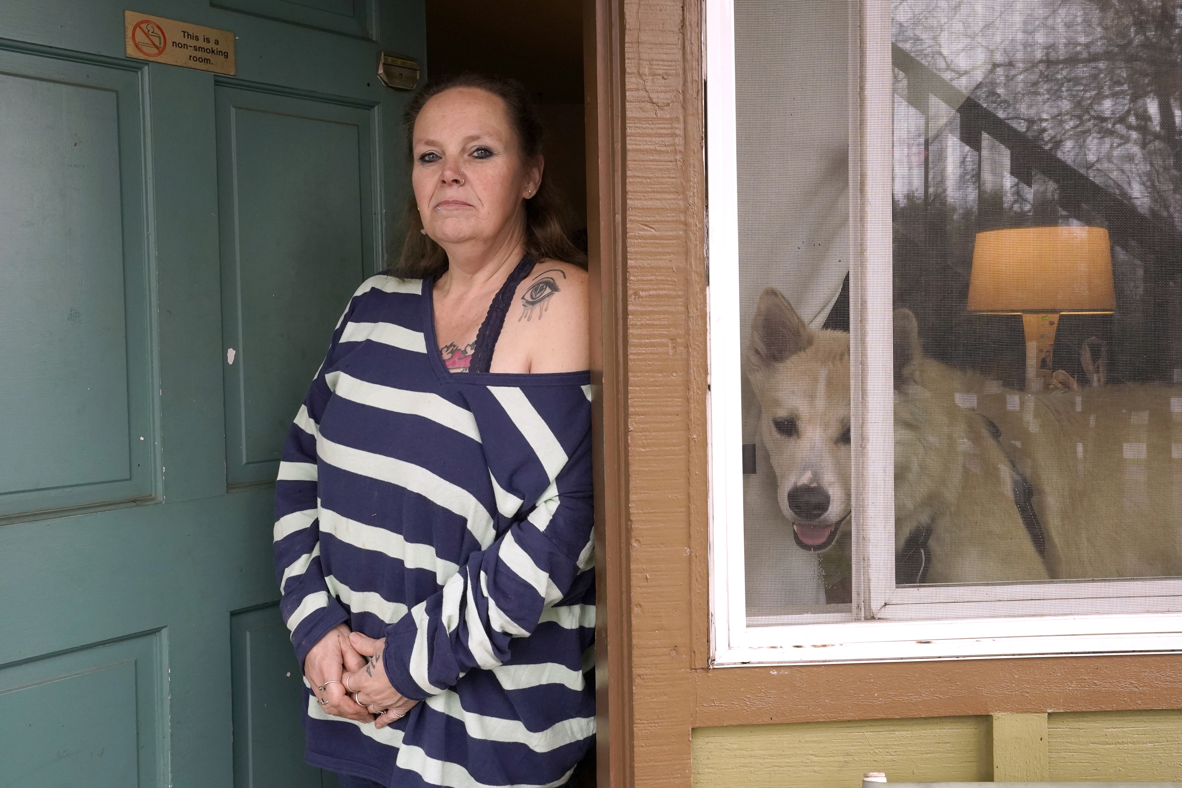 Stephanie Lammers stands in the front door of her house and looks directly towards the camera. Her dog is visible in the window beside her.