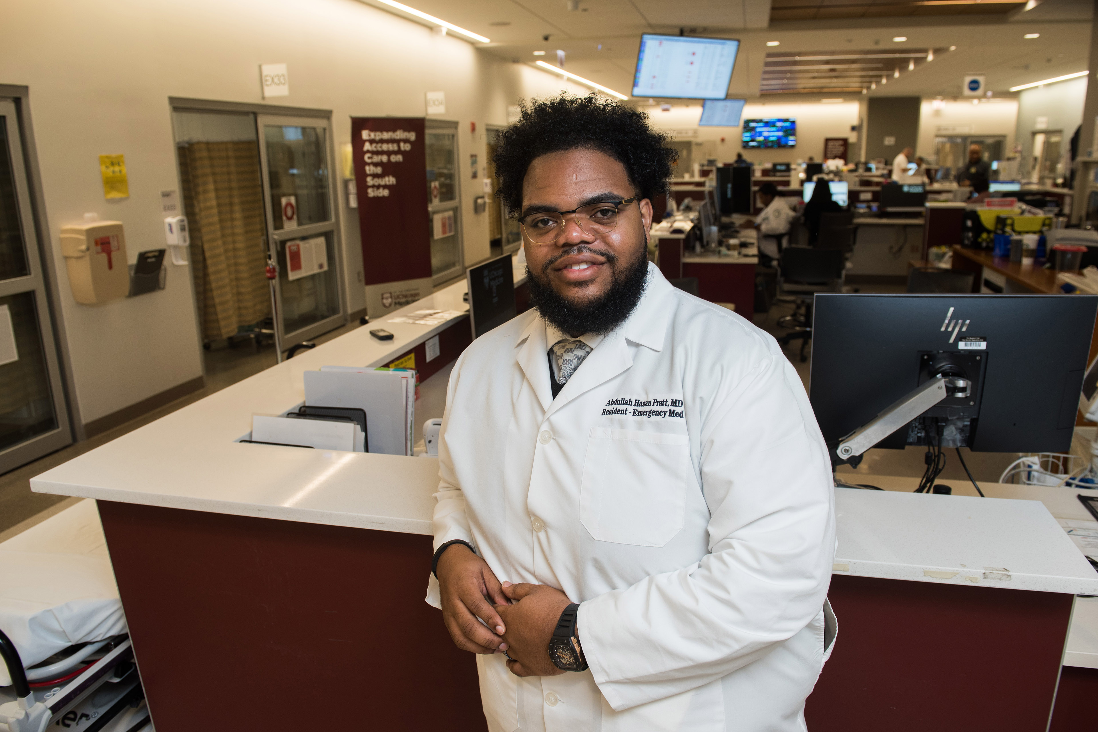 Dr. Abdullah Pratt stands at a reception desk in a medical building. He wears a white doctor's coat and gently smiles at the camera.