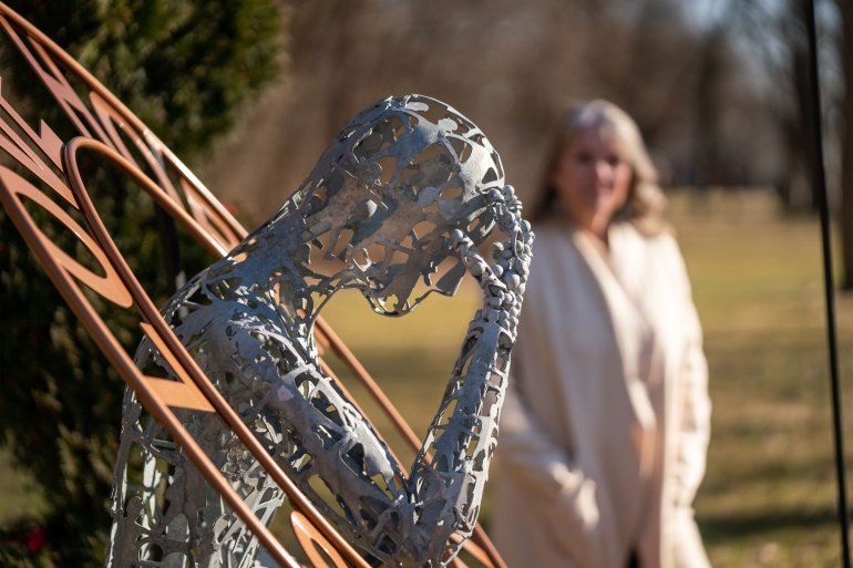 A woman in a white coat, out of focus in the background of the image, looks at a statue in a park. The metal statue, in the foreground, is abstract but appears to be a person leaning their head on their hand and surrounded by a light brown ring of words.