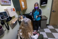 One woman in a mask facing the camera speaks to another woman facing away from the camera in a clinical office.