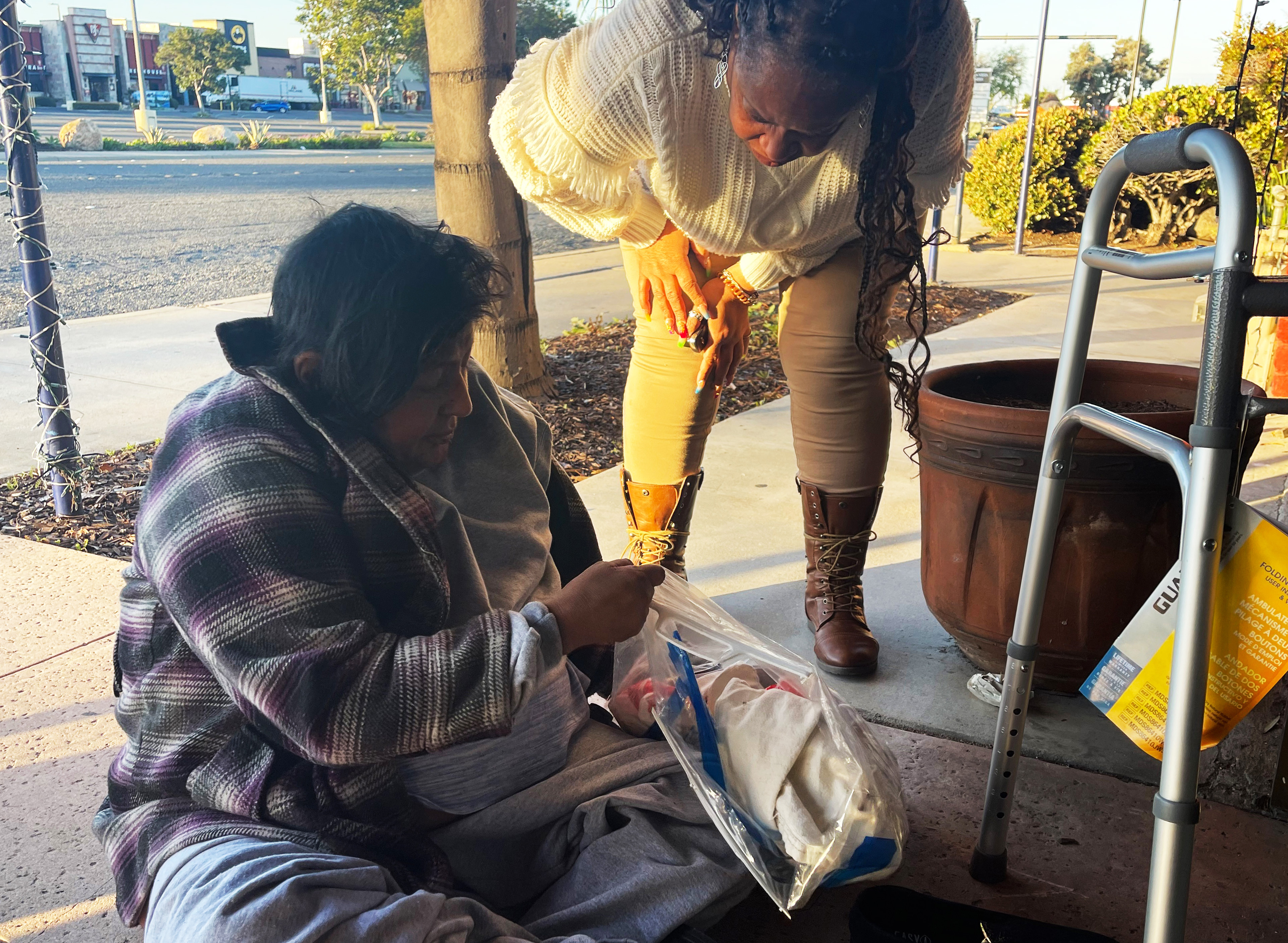 A photo of a homeless woman sitting and looking into a bag of supplies. Another woman is standing and watching her.