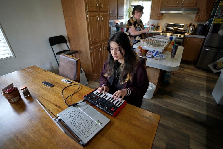 A photo of teenager using her keyboard and laptop to make music while her mother cooks in the kitchen behind her.