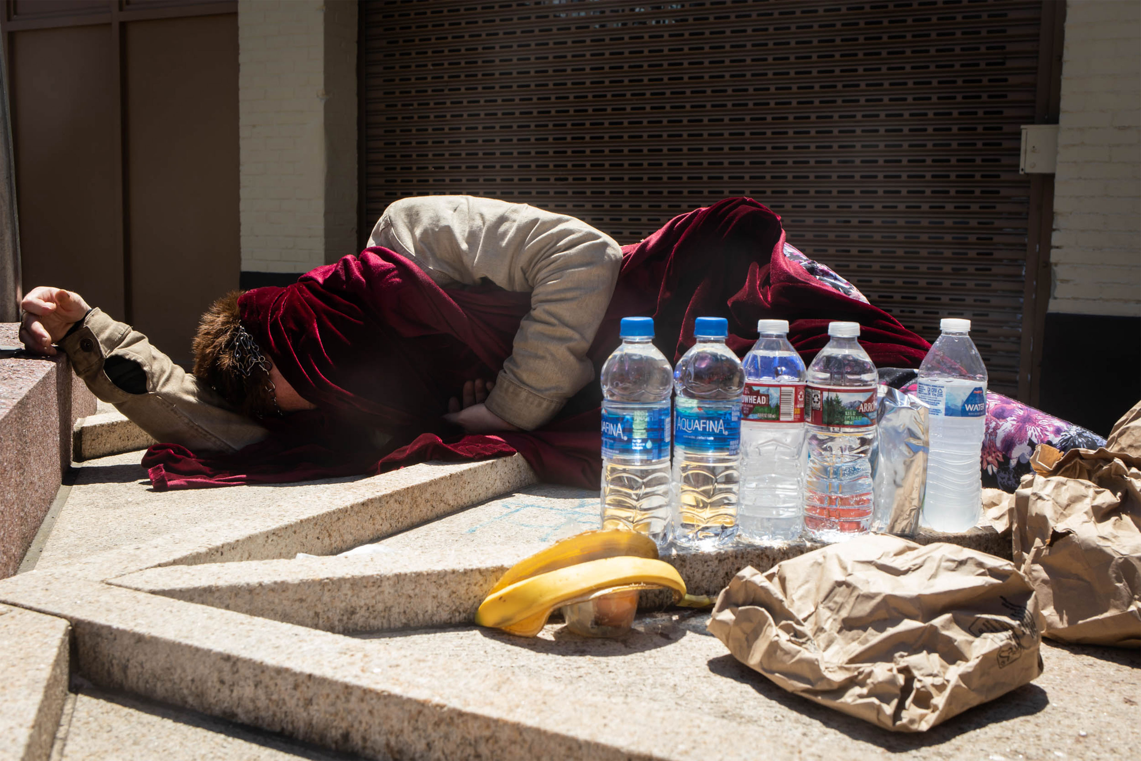 A photo of a person lying down on the ground with a blanket covering them next to water bottles and bags of food.