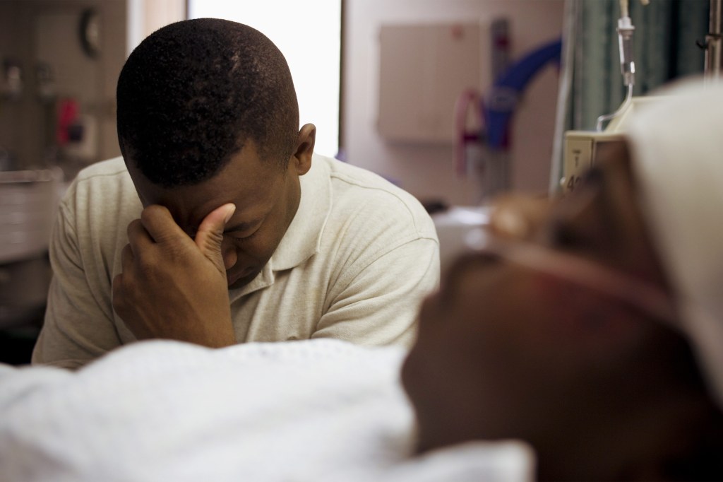 A photo of a Black man sitting beside his son in a hospital bed, covering his face.