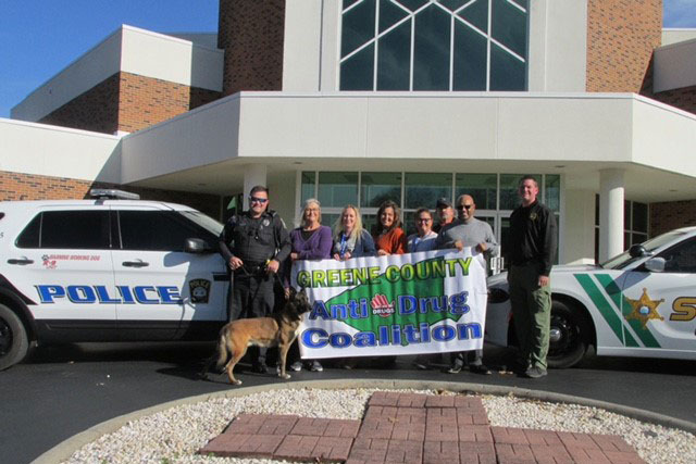 Nine people and a dog stand in front of a building and two police vehicles. Some of the people hold a banner that reads "Greene County Anti-Drug Coalition".